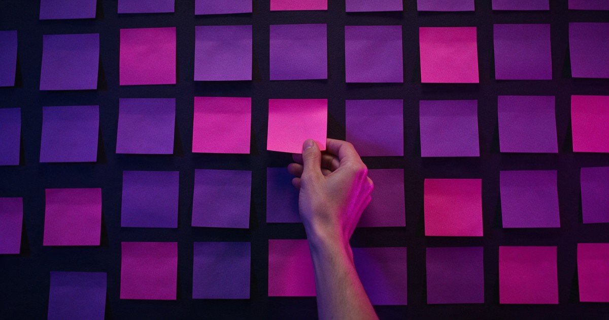 Mão cola um papel adesivo magenta em uma parede coberta de papéis adesivos em branco.